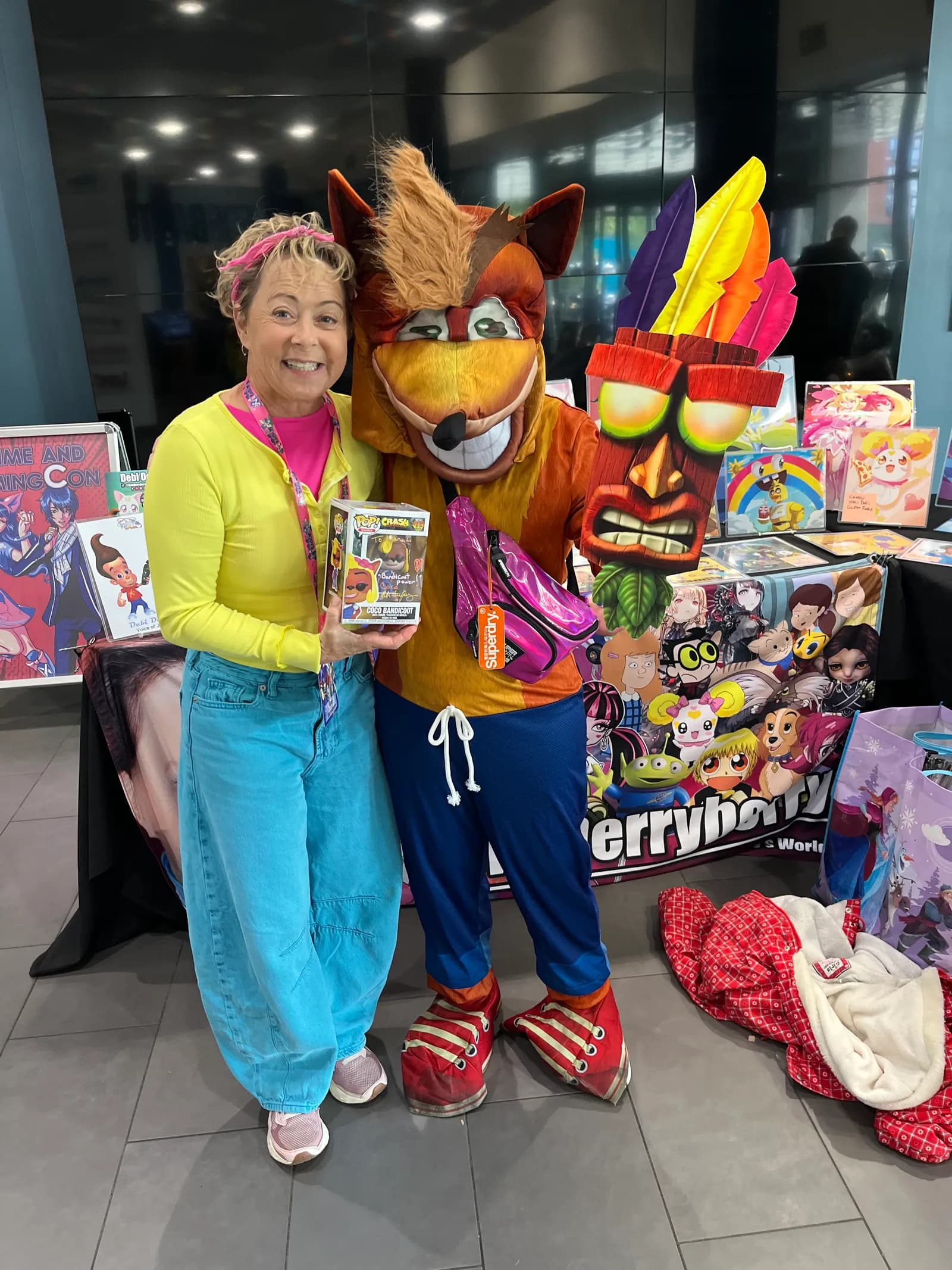 A smiling woman in bright clothing stands next to a Crash Bandicoot mascot, holding a Crash Bandicoot Funko Pop! figurine, with colorful merchandise in the background.