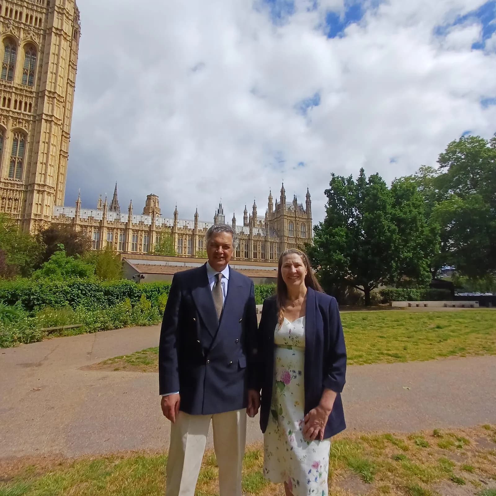 A man in a suit and a woman in a floral dress stand in a park with the Houses of Parliament in the background under a cloudy sky.