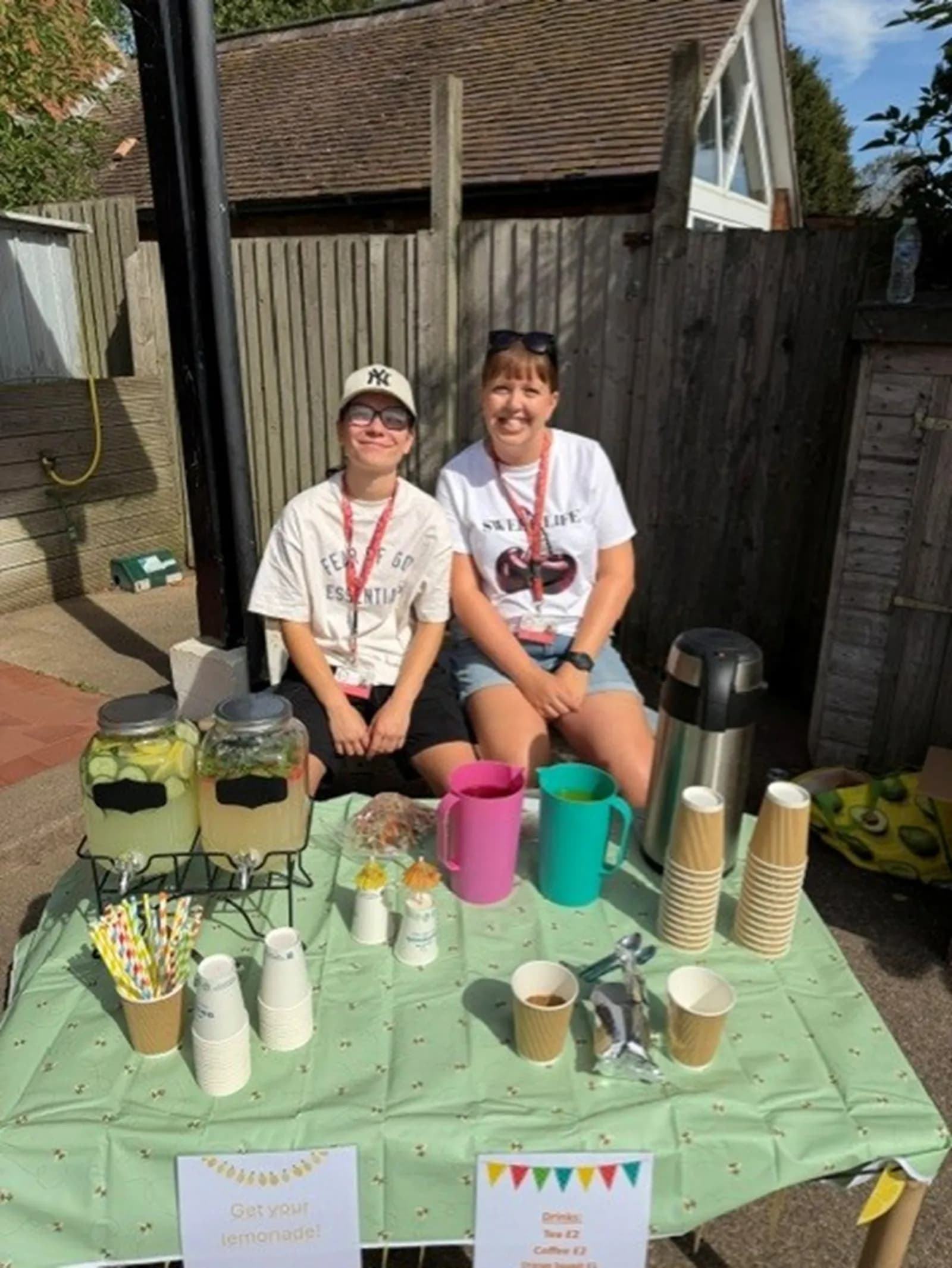 Two women smile while sitting behind a table with two lemonade dispensers and other drinks set up for sale.