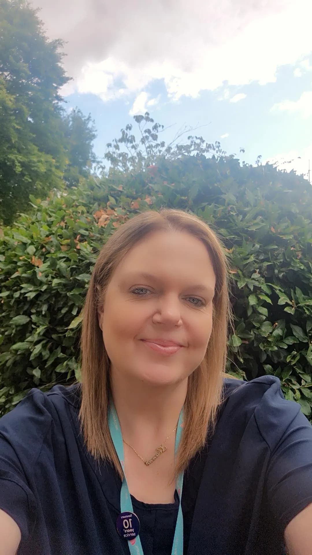 A woman with light brown hair smiles while looking directly at the camera, with green foliage and a partly cloudy sky in the background.